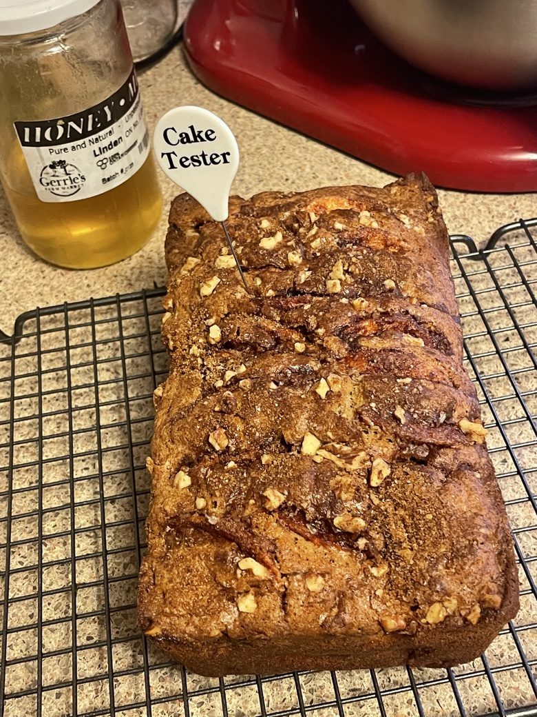 Apple walnut loaf studded with walnuts on a cooling rack, with a cake tester and honey jar