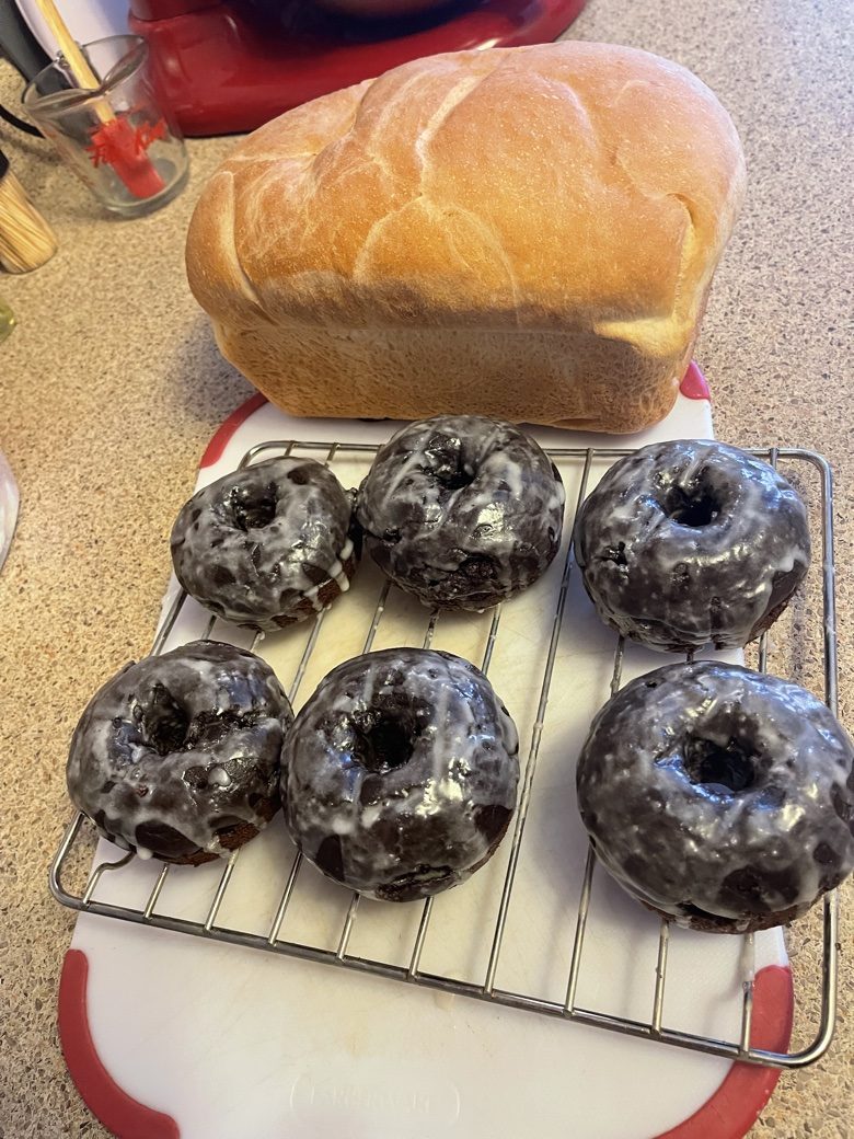 Six glazed chocolate donuts on a cooling rack with a fresh loaf behind them